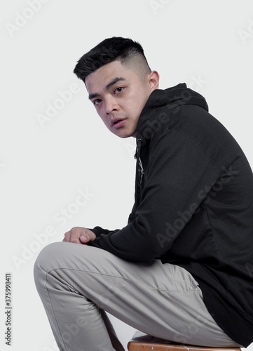 Young handsome wearing black zip hoodie is sitting on a chair isolated on background © DendraCreative