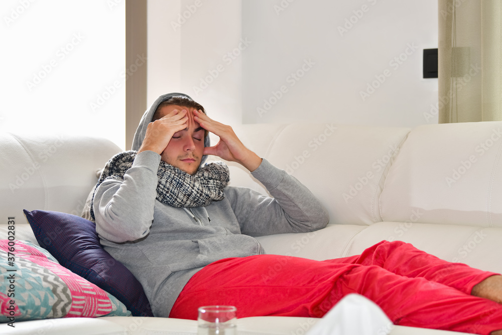 Young man looking in pain lying on a sofa while holding his head with both hands. Sickness and healthcare concept.