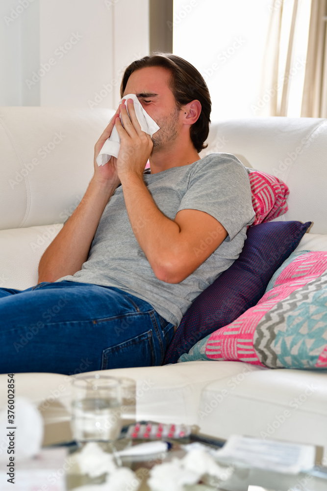 Young man blowing his nose in a napkin while lying on a sofa on an out of focus background. Hygiene and relaxation concept. Young man blowing his nose in a napkin while lying on a sofa on an out of focus background. Hygiene and relaxation concept.