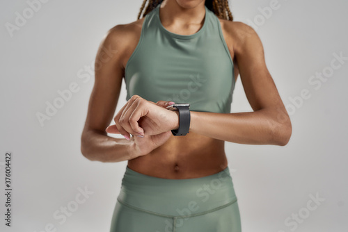 Close up shot of a athletic mixed race woman setting up her smartwatch or checking fitness results while standing isolated over grey background