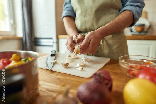 Close up of hands of man in apron peeling garlic while preparing healthy meal, soup in the kitchen. Cooking at home, Italian cuisine concept