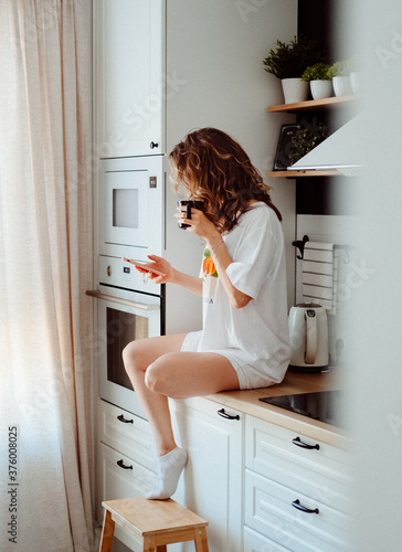 Young pretty woman drinks coffee sitting at her kitchen at home and using smartphone enjoys of morning. Smiling woman reading phone message.