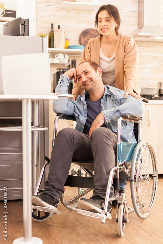 Positive man in wheelchair and wife looking at laptop in kitchen. Disabled paralyzed handicapped man with walking disability integrating after an accident.