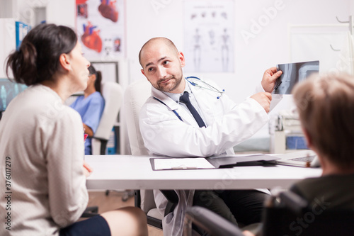 Doctor in hospital office explaining knee injury on x-ray image to young woman and her disabled mother in wheelchair. Medic in white coat with stethoscope discussing diagnosis with patient. © DC Studio