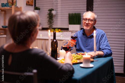 Senior man telling a story to his wife while celebrating in kitchen with wine and food. Senior couple sitting at the table in dining room   talking  enjoying the meal  celebrating their anniversary in