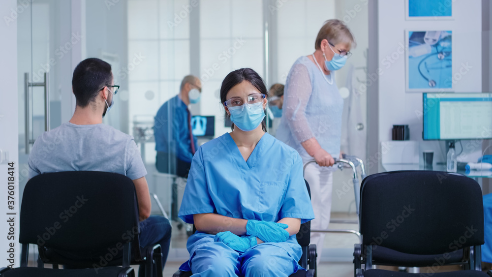 Obraz premium Worried nurse with face mask against coronavirus sitting on chairs in hospital waiting room. Disabled senior woman with walking frame in hospital hallway.