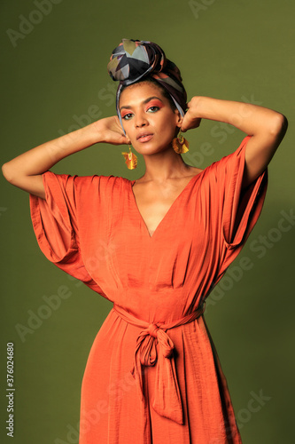 Fashion studio photo of stylish african woman with turban and orange dress posing over green background.