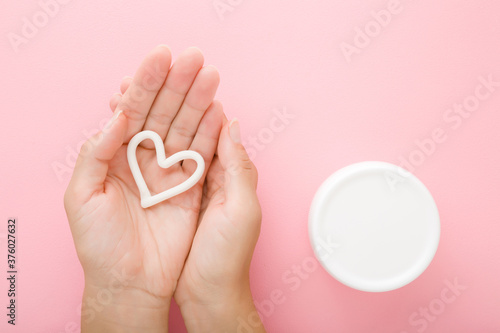 Heart shape on young woman palm. White cream jar on light pink table background. Pastel color. Care about clean and soft body skin. Closeup. Point of view shot. Top down view.