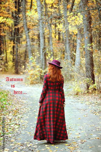 Happy young woman with red hair and freckles in dress, hat walking in yellow autumn park at nature. Fall season