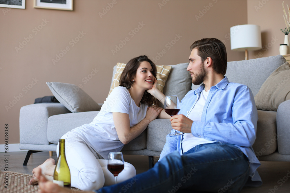 Happy couple sitting, relaxing on floor in living room, drinking red wine. Smiling young husband and wife rest at home enjoy romantic date on family weekend together