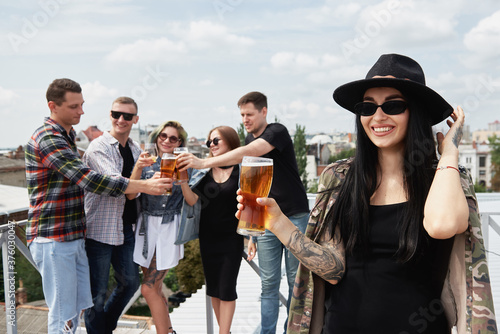 Group of friends drinking beer before festival at outdoors pub on roof, toasting and laughing, copy space. Friendship and celebration concept