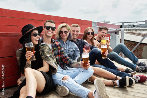 Group of friends drinking beer before festival at outdoors pub on roof, toasting and laughing, copy space. Friendship and celebration concept