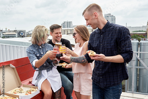 Group of friends drinking beer and eating pizza before festival at outdoors pub on roof, toasting and laughing, copy space. Friendship and celebration concept photo