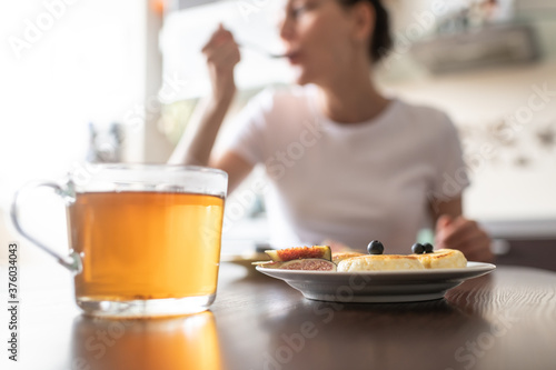 Unfocused woman having breakfast at home. Focus is on the plate with cheese pancakes