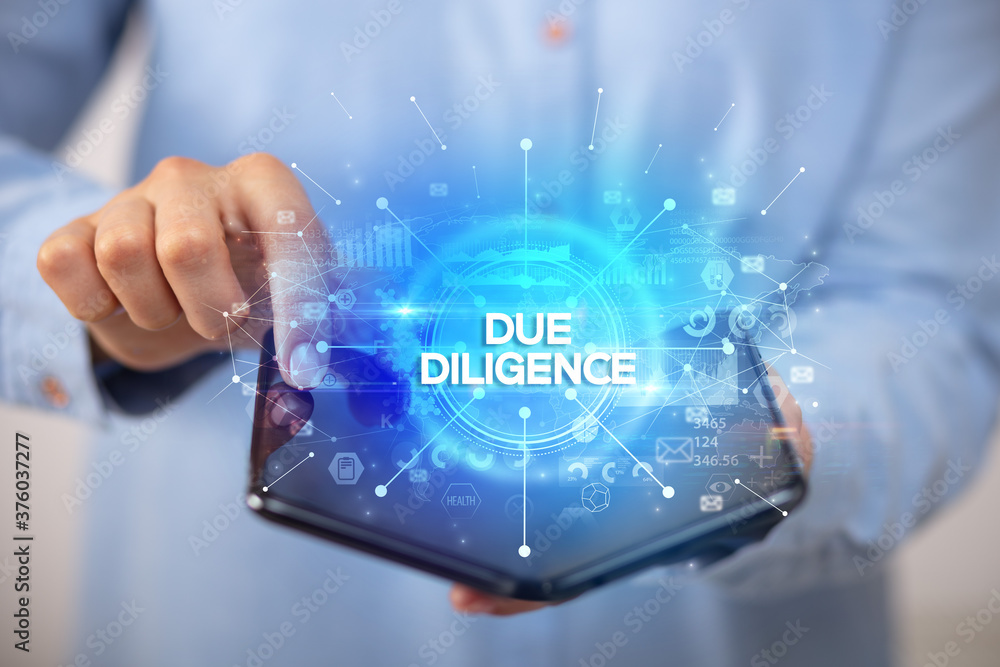 Businessman holding a foldable smartphone with DUE DILIGENCE inscription, new business concept Businessman holding a foldable smartphone with DUE DILIGENCE inscription, new business concept