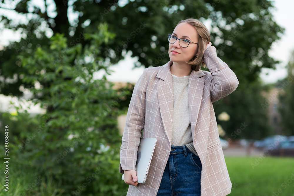 A girl of European appearance in glasses is a student with a laptop in the fresh air.A woman in office style stands in the Park with a computer, and adjusts her hair.the concept of remote work A girl of European appearance in glasses is a student with a laptop in the fresh air.A woman in office style stands in the Park with a computer, and adjusts her hair.the concept of remote work