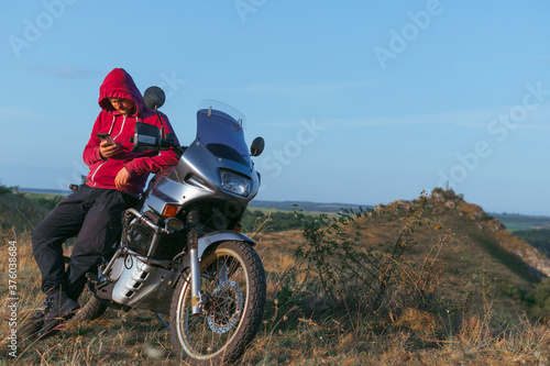 A man in a red sweatshirt with a hood. Motorcycle driver at the top of the hill overlooking the rocks and the river. Hobbies and leisure. Far from civilization.