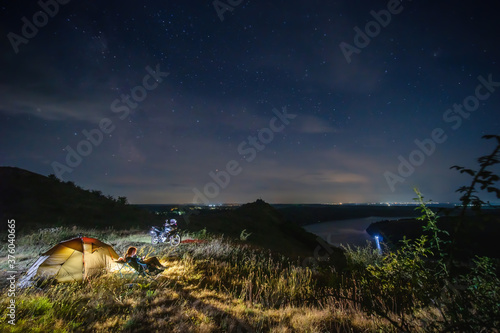 Night camp on the top of the mountain, Camping with tent and motorcycle, Woman is sitting on folding chairs. Starry sky with the milky way. Starfall. Concept of active lifestyle and tourism equipment © Sergey