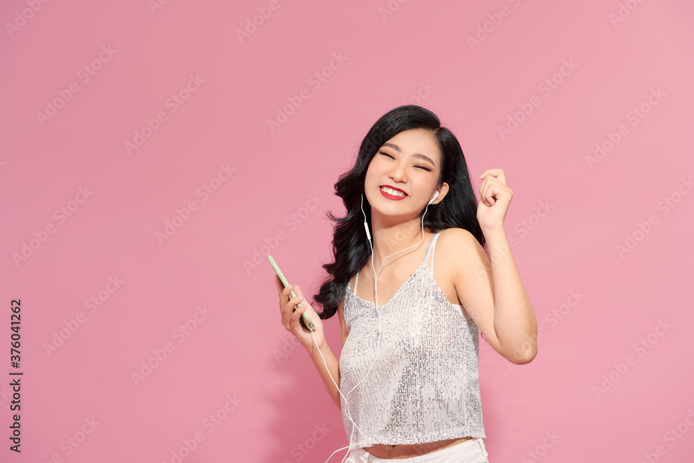 Cute young female in modern headphones cheerfully smiling and looking up while standing on pink background and listening to music Cute young female in modern headphones cheerfully smiling and looking up while standing on pink background and listening to music