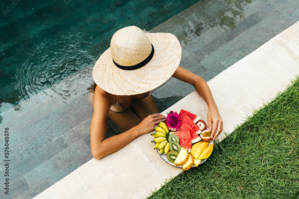 Girl relaxing and eating fruits near the pool on luxury villa in Bali. Exotic summer diet. Tropical beach lifestyle.