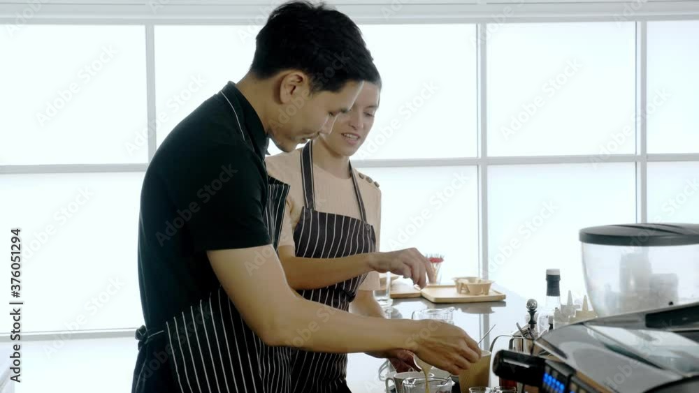 Multi ethinic colleague working together in cafe concept. Hand held, medium shot of male asian barista preparing coffee while female caucasian colleage teaching, giving comment and support Multi ethinic colleague working together in cafe concept. Hand held, medium shot of male asian barista preparing coffee while female caucasian colleage teaching, giving comment and support