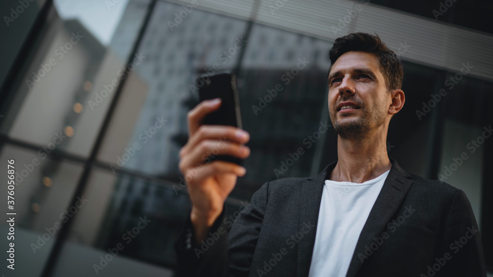 Happy confident businessman texting with client via smartphone discussing new sustainable project, Young male manager using cellphone outdoor near modern business center building Happy confident businessman texting with client via smartphone discussing new sustainable project, Young male manager using cellphone outdoor near modern business center building