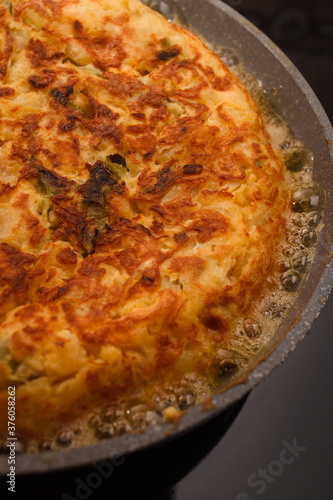 Tortilla de Patatas in a pan with eggs, potatoes, onion and green peppers on a pan. Traditional spanish dish. Rustic black background.
