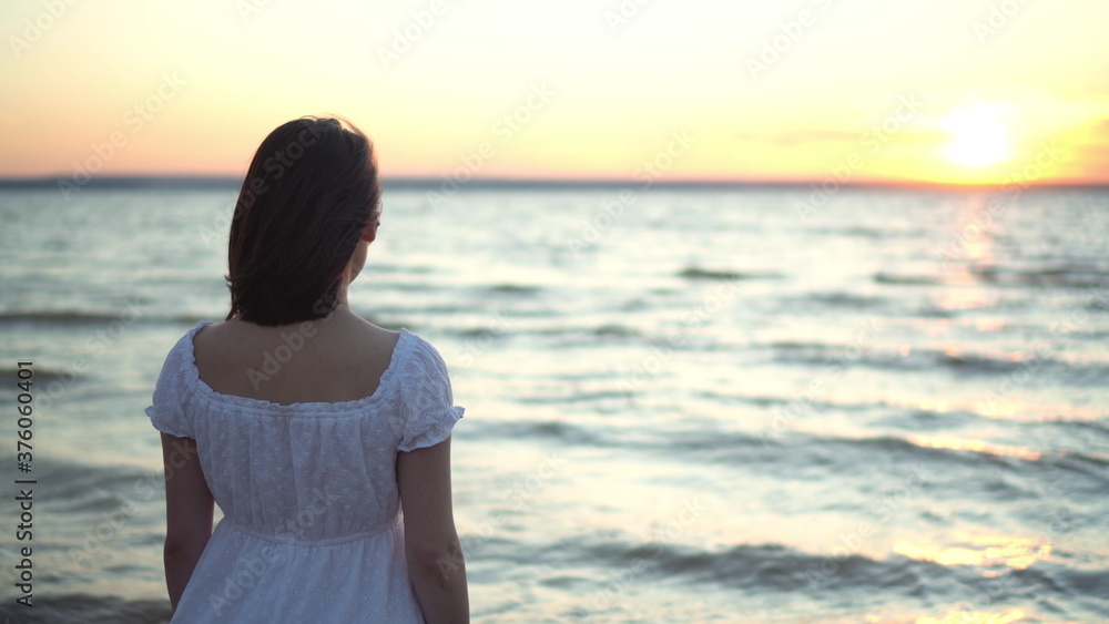 Attractive young woman looks at the sunset on the beach by the sea. The girl in a white dress stands with her back. Attractive young woman looks at the sunset on the beach by the sea. The girl in a white dress stands with her back.