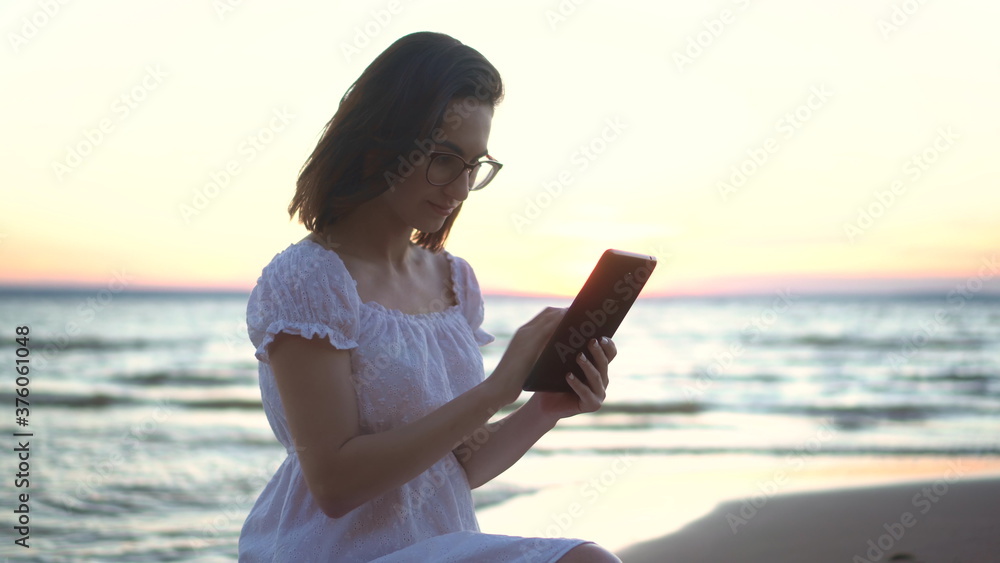 A young woman sits on a stone on the beach by the sea with a tablet in her hands. A girl in a white dress at sunset is chatting on a tablet closeup.