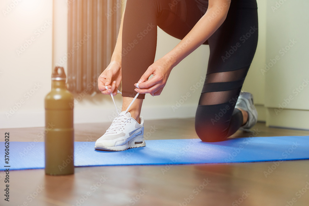 A fitness girl in a black workout tight suit who is tying her shoelaces on white sneakers while kneeling on the blue yoga mat near a green aluminum bottle of water before training at home. A fitness girl in a black workout tight suit who is tying her shoelaces on white sneakers while kneeling on the blue yoga mat near a green aluminum bottle of water before training at home.