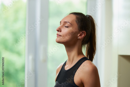A side view photo of a sporty slim girl with closed eyes in a black workout tight suit is relaxing before training near the window at home. Free Download A side view photo of a sporty slim girl with closed eyes in a black workout tight suit is relaxing before training near the window at home. - GFXLoad