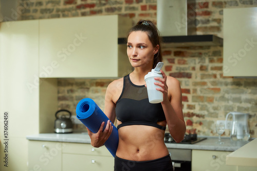 A smiling sporty girl in a black workout tight suit is holding the blue yoga mat and white plastic shaker to quench her thirst after training at home. © Roman Tyukin