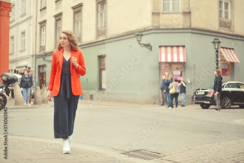 Stylish hipster girl posing in front of the old street in city. Trendy young woman model. Street fashion concept, wearing  red jacket. Tourist smile, sunny day. Elegant girl with long curly hair © MoreThanProd