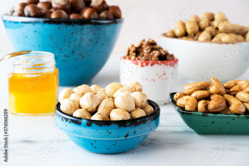 Different nuts (hazelnuts, cashews, walnuts), peeled and in shell in bowls of different colors (blue, white, green) stand on a white background. There is a small jar of honey nearby. Close-up