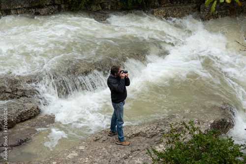 A swollen torrent cascades down the mountain between rocks. A man stands by the water and takes pictures.