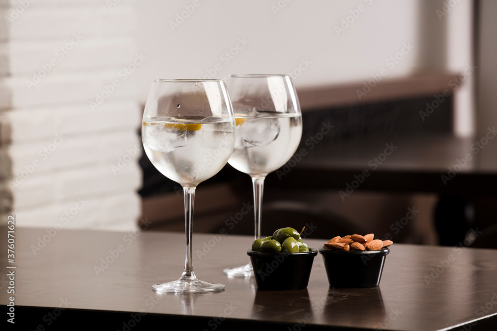 large glasses with alcoholic drink, ice and a piece of natural orange and container with olives and almonds, on top of the wooden table, inside the restaurant and free space for decoration.