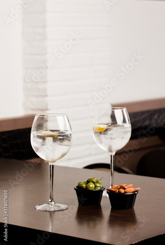 large glasses with alcoholic drink  ice and a piece of natural orange and container with olives and almonds  on top of the wooden table  inside the restaurant and free space for decoration.