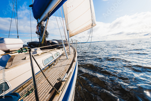 Sloop rigged modern yacht with wooden teak deck sailing near the rocky shores of Tarbert after the rain. Scotland, UK. Close-up view from the deck to the bow, mast and sails. Clear blue sky photo