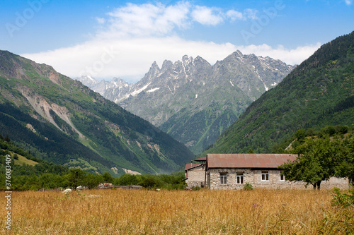 Amazing mountain landscape in Svaneti, Georgia on a sunny day. At the foot of a single-story building and field with yellow plants. Magnificent Caucasus.