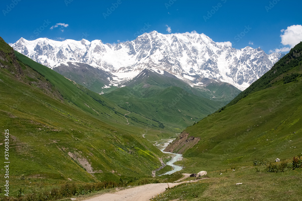 Beautiful mountain landscape in Georgia Svaneti. The snow-capped peaks of mount Shkhara near the Ushguli village are visible on a sunny day. A river flows between the green hills
