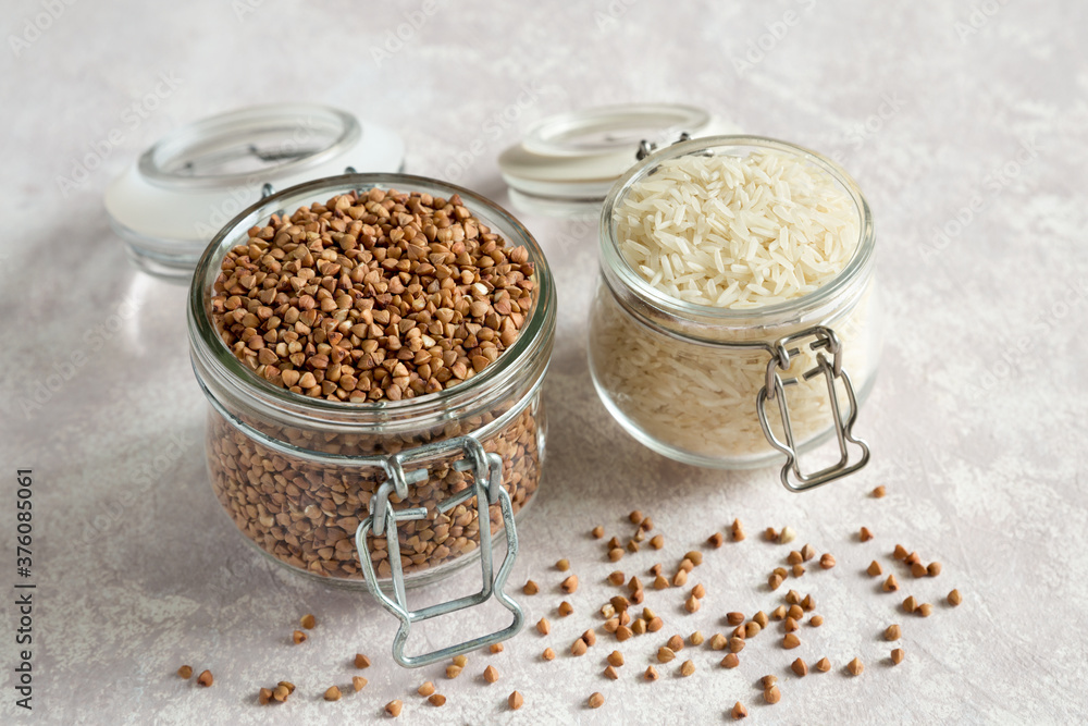 Two glass jars with uncooked rice and buckwheat grains close up on a light background. Storage of cereals. Healthy food. Storage organic products Two glass jars with uncooked rice and buckwheat grains close up on a light background. Storage of cereals. Healthy food. Storage organic products