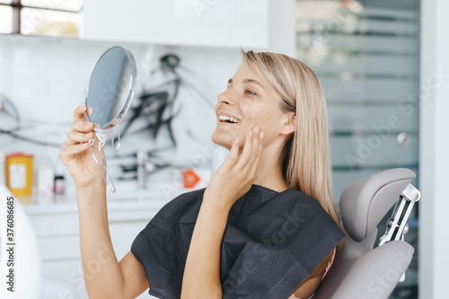 Young woman patient is looking in the mirror and admires her new smile after dental treatment in the dentistry clinic.