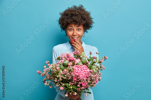 Women, greeting and celebration concept. Positive Afro American woman holds beautiful bouquet of different flowers stands against blue background. Glad mom gets present from daughter on spring holiday