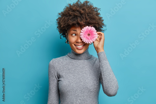 Portrait of happy enrgetic ethnic girl has curly hair covers eye with gerbera flower, smiles positively, wears casual turtleneck, going to make bouquet or decorate room, blue background, has fun © Wayhome Studio
