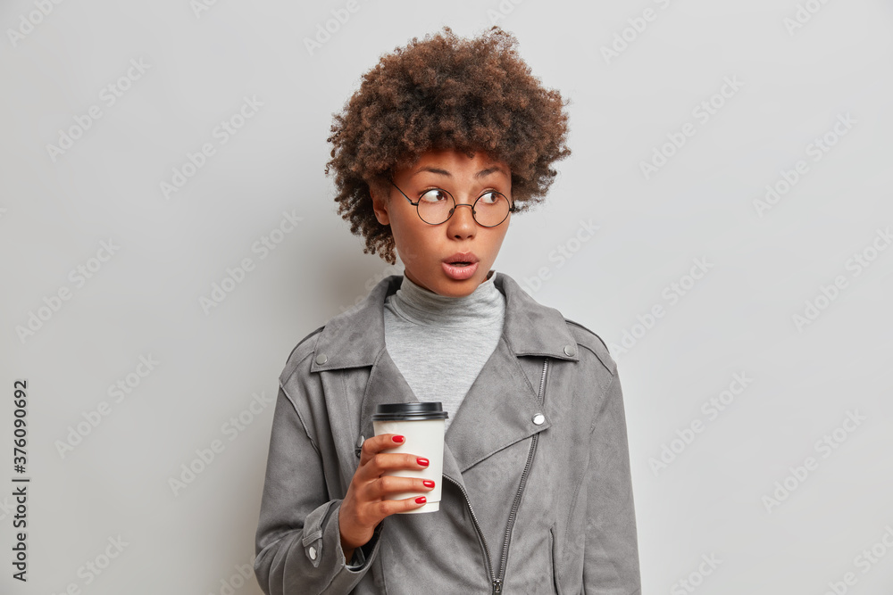Isolated shot of surprised young African American woman holds disposable cup of coffee, notices something astonishing aside, wears optical glasses, grey jacket, isolated on white background. Isolated shot of surprised young African American woman holds disposable cup of coffee, notices something astonishing aside, wears optical glasses, grey jacket, isolated on white background.