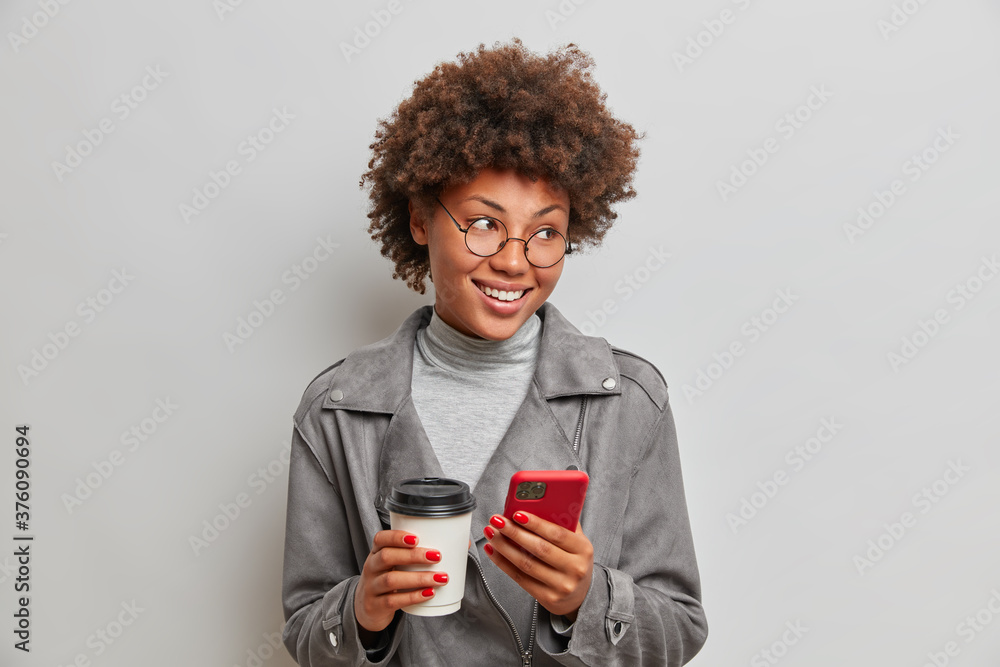 Positive good looking student has coffee break, holds mobile phone and disposable cup in hands, looks away gladfully, dressed in stylish grey jacket, isolated on white background. Lifestyle concept Positive good looking student has coffee break, holds mobile phone and disposable cup in hands, looks away gladfully, dressed in stylish grey jacket, isolated on white background. Lifestyle concept
