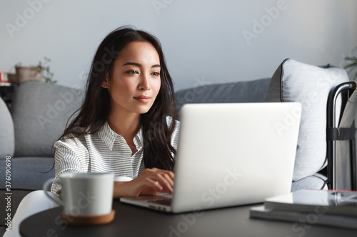 Young ambitious asian girl working remote from home, looking at laptop screen and smiling. Woman checking mail or researching while telecommuting, sitting on floor at her apartment photo
