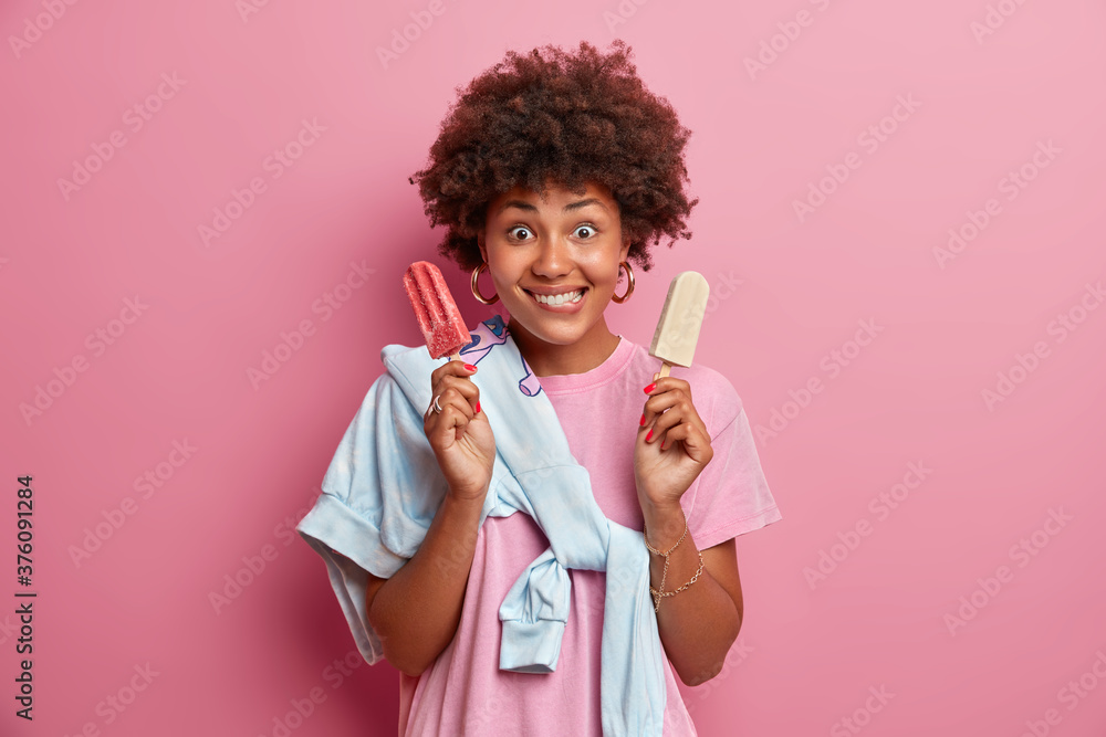 Beautiful cheerfull millennial girl poses with delicious cold popsicle, bites lips, invites friend to have walk and eat summer dessert, wears t shirt, sweater tied over shoulder, poses indoor Beautiful cheerfull millennial girl poses with delicious cold popsicle, bites lips, invites friend to have walk and eat summer dessert, wears t shirt, sweater tied over shoulder, poses indoor