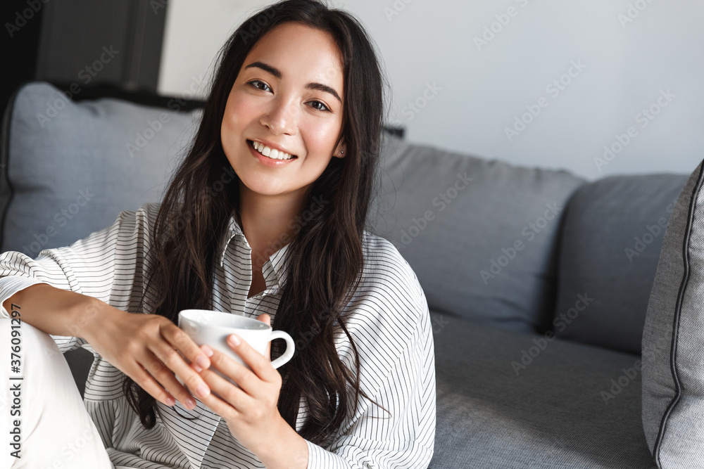 Close-up of beautiful brunette asian girl sitting at home and smiling, drinking morning cup of coffee. Woman enjoying tea and watching tv, relaxing in her apartment, having lazy weekend Close-up of beautiful brunette asian girl sitting at home and smiling, drinking morning cup of coffee. Woman enjoying tea and watching tv, relaxing in her apartment, having lazy weekend