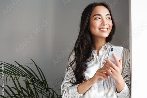 Portrait of attractive modern asian woman looking outside window and smiling dreamy, holding smartphone. Girl waiting for phone call, working remote from home, planning schedule photo